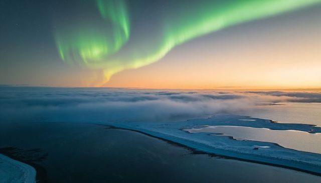Dancing green aurora over arctic coastline at twilight with snow, fog and icy water