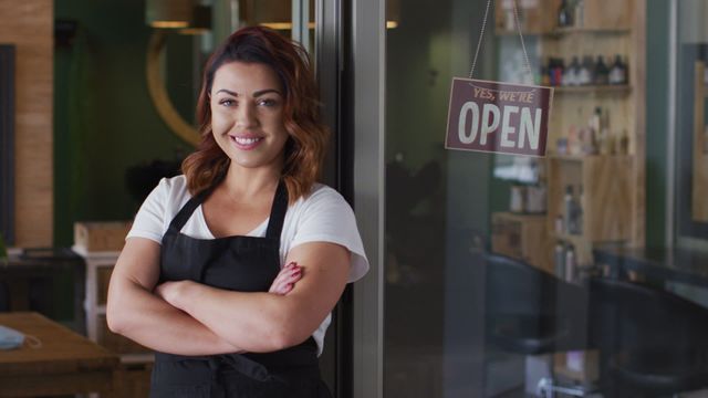 Female hairdresser posing confidently with arms crossed in beauty salon doorway with open sign in view. Perfect depiction of a friendly and professional business owner ready to welcome clients. Ideal for use in articles or advertisements promoting small businesses, entrepreneurship, or the hairdressing industry.