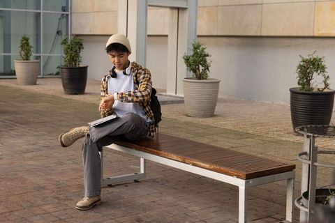 Asian teen checking smartwatch on bench with notebook and backpack wearing headphones
