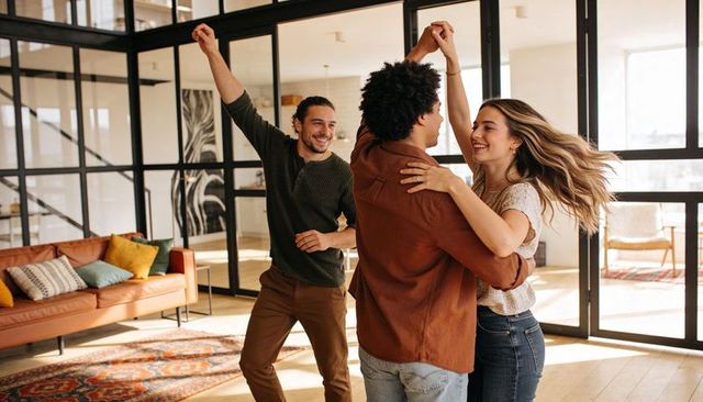 Young couple dancing in sunlit modern apartment with friend celebrating lively home moment