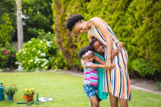 This image depicts a joyful moment of an African American mother embracing her two daughters in a lush backyard. Ideal for use in family-oriented advertisements, lifestyle blogs, and articles about parenting, outdoor activities, and family bonding. The vibrant greenery and casual setting make it perfect for promoting outdoor products, gardening tools, and summer activities.