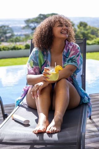 African american woman laughing and relaxing poolside, sipping citrus drink, sunscreen nearby