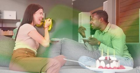 Couple sharing birthday cake on sofa laughing and feeding each other in cozy living room
