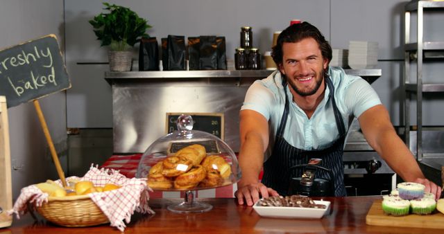 This image features a cheerful baker standing behind a wooden counter full of freshly baked pastries inside a modern bakery cafe. A variety of breads, cookies, and cupcakes are attractively displayed. The friendly and welcoming atmosphere is ideal for promoting a local bakery, highlighting artisanal baking, or showcasing bakery products. Perfect for use in marketing materials, social media posts, or culinary blogs emphasizing handmade and freshly baked goods.