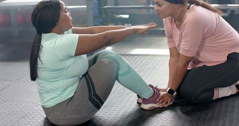 Diverse women doing sit-ups while partner holds feet on gym floor in activewear