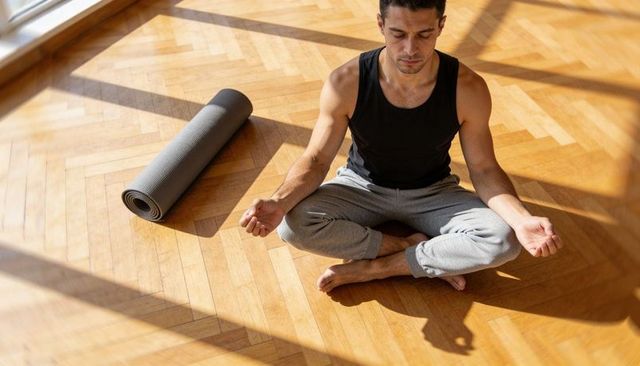 Sunlit man meditating cross-legged on parquet floor with rolled yoga mat nearby at home