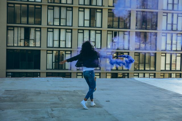 Rear view of a hip young biracial woman wearing leather jacket and jeans, holding a smoke grenade with purple smoke on an urban rooftop with building in the background