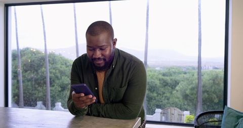African American man leaning on kitchen counter using smartphone by large window wearing earbud