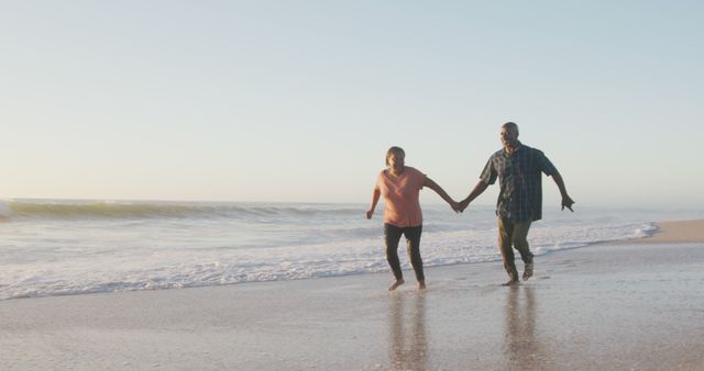 Smiling senior african american couple holding hands and running on sunny beach. healthy, active retirement beach holiday.