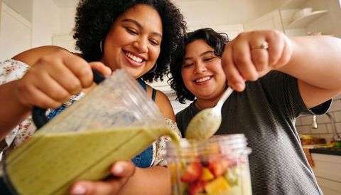 Smiling friends pouring green smoothie over fresh fruit into jar in bright kitchen