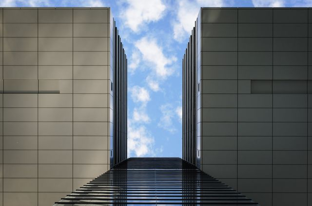 This image captures the view upwards between two modern high-rise buildings with a blue sky background. The geometric symmetry and sleek design emphasize urban innovation and architectural progress. Ideal for use in marketing materials for real estate companies, architectural firms, and city development projects.