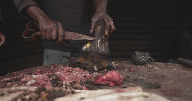 Butcher is cutting a goat's head with a large knife on a table covered in meat and blood. Dark lighting creates a rustic, traditional atmosphere. Suitable for topics related to traditional butchery, food preparation, meat processing, cultural practices, or restaurant kitchen activities.