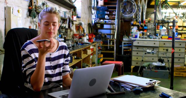 Young woman using voice command on smartphone while working at a creative workshop. Suitable for themes related to technology, engineering, creativity, modern workspace, remote work, and industrial environments.