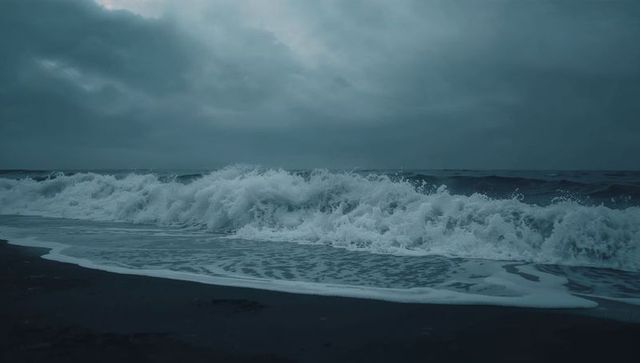 Crashing waves rolling onto dark sand beach under moody overcast sky