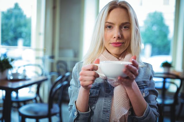 Blonde woman sipping coffee in cozy cafe, wearing casual denim jacket and scarf. Perfect for concepts of relaxation, casual lifestyle, coffee culture, and indoor leisure.