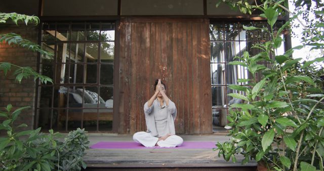 Woman sitting on yoga mat in cross-legged position, performing meditation outside house surrounded by lush greenery. Serenity and peaceful ambiance make it suitable for wellness blogs, meditation guides, outdoor retreat promotions, and lifestyle publications focused on mindfulness and relaxation.
