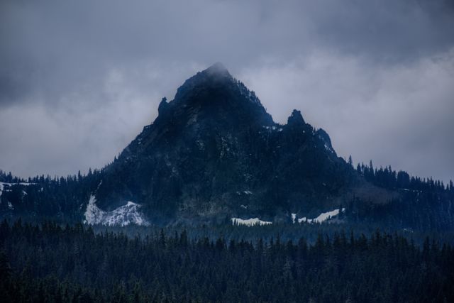 Dramatic view of a cloudy mountain peak surrounded by a forest with patches of snow. The sky is foreboding, adding a sense of mystery and adventure. Ideal for use in travel and outdoor adventure content, nature documentaries, weather features, and atmospheric landscape visuals.
