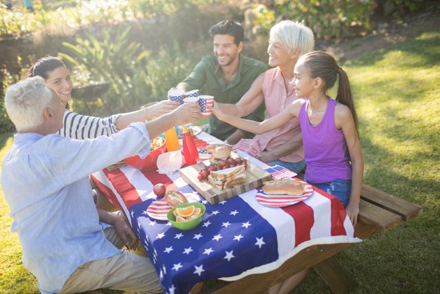 Family enjoying a picnic in the park on a sunny day, using an American flag tablecloth. They are toasting with cups and sharing a meal, creating a joyful and bonding experience. Perfect for illustrating family gatherings, outdoor celebrations, and patriotic events.