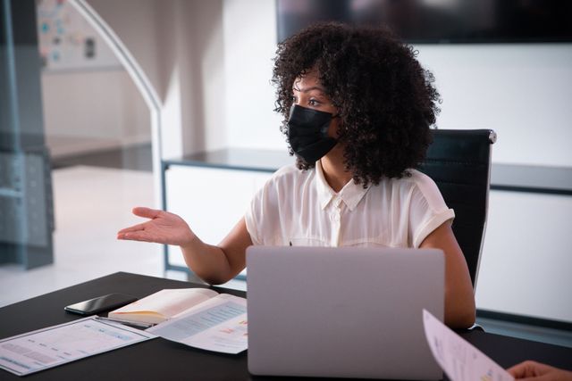 African american businesswoman wearing face mask in discussion, sitting at desk with laptop. working in business at a modern office during coronavirus covid 19 pandemic.