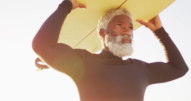 Senior african american man walking with surfboard on sunny beach. healthy and active time beach holiday.