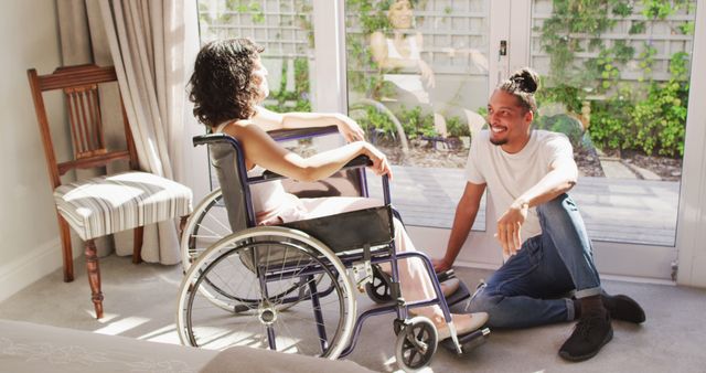 Happy biracial woman in wheelchair and smiling male partner talking in sunny living room. wellbeing and domestic lifestyle with physical disability.