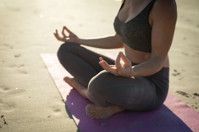 This image depicts a woman practicing yoga on a beach at sunrise. She is sitting on a yoga mat in a meditative pose, focusing on relaxation and mindfulness. Ideal for use in wellness blogs, fitness websites, meditation guides, and promotional materials for yoga retreats or outdoor fitness programs.