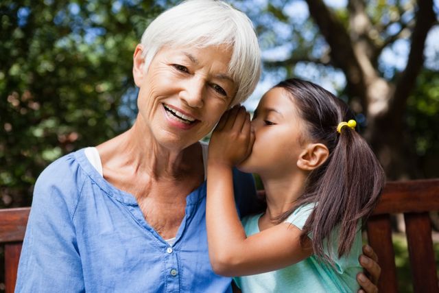 Young girl whispering secret to smiling grandmother, who is enjoying the moment. Ideal for themes of family bonding, multi-generational interactions, and outdoor activities. Suitable for promoting family products, retirement planning, or lifestyle blogs.