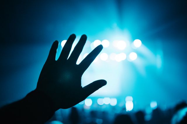 Silhouette of hand raised at a concert, set against vibrant blue stage lights. Great for themes related to live music, entertainment, nightlife, and the energy of concerts. Can be used in promotional material for events, wallpapers for musicians, and backgrounds for social media posts.