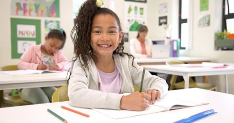 Smiling Schoolgirl Writing in Classroom with Educational Posters