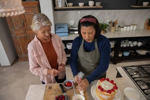 Two women decorating strawberry layer cake with piping bag in modern home kitchen