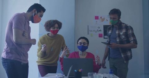 Diverse team collaborating around laptop and tablet while wearing face masks in office