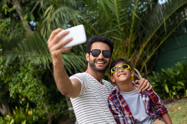Father and son enjoying quality time together in a yard, both wearing sunglasses and smiling while taking a selfie. Ideal for use in family-oriented advertisements, social media posts about family bonding, summer activities, or outdoor leisure. The tropical background with palm trees adds a vacation vibe, making it suitable for travel promotions or lifestyle blogs.