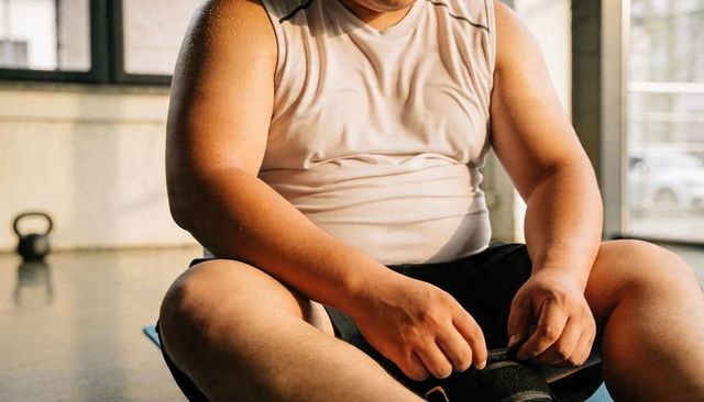 Overweight man adjusting ankle strap on mat after intense workout sweating in sunlit gym
