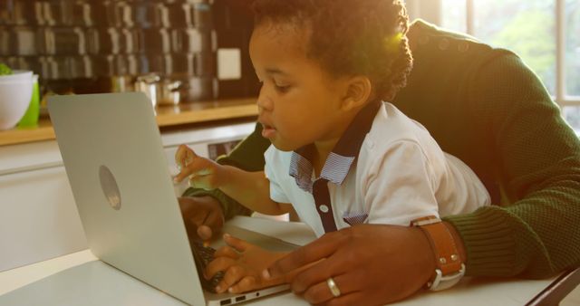 Father and young son are utilizing a laptop together in a sunlit kitchen, creating a warm and engaging atmosphere. This appealing image could be used for family-oriented advertising, articles on parenting, technology use in education, or home living. It effectively showcases the bond and interaction between father and child while embracing modern technology in a cozy home environment.