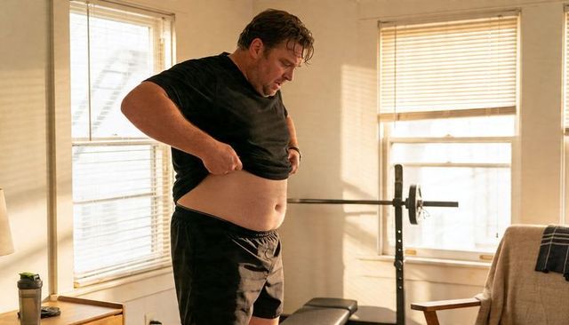 Man pulling up shirt and checking abdomen in sunlit home gym during casual workout routine