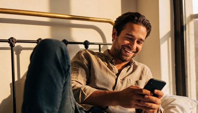 Man Relaxing on Bed Laughing at Smartphone in Sunlit Cozy Bedroom