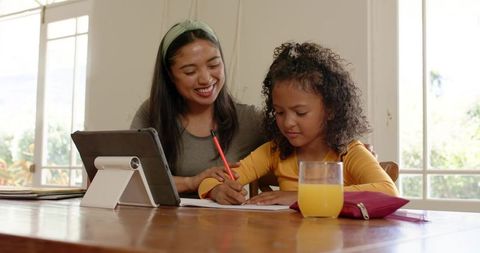 Parent guiding child with tablet while doing homework at sunlit wooden kitchen table