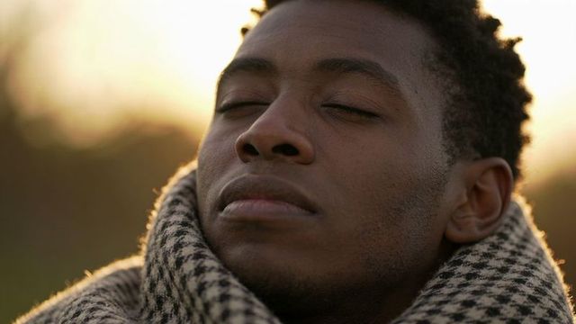 African american man closing eyes, tilting head upward at golden hour wearing wool scarf