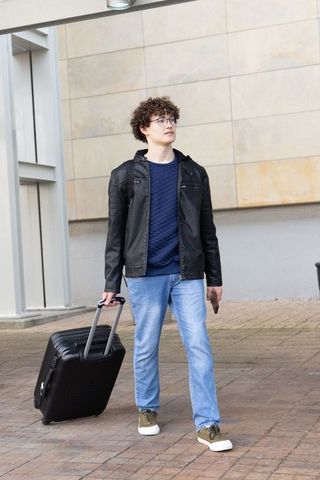Young traveler walking pulling wheeled suitcase checking smartphone in urban plaza