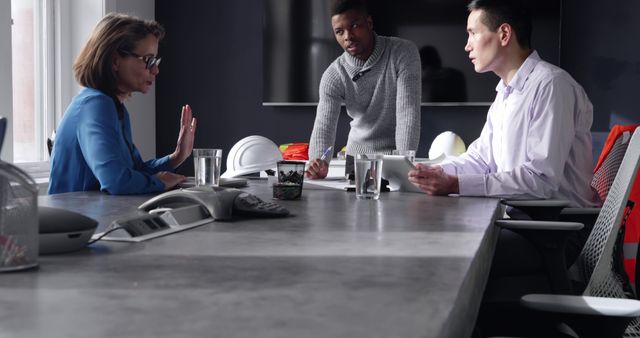 Three colleagues engaged in a serious discussion around a conference table in a modern office environment. This image conveys themes of teamwork, collaboration, and communication, making it suitable for business-related contexts, corporate websites, and presentations focused on work culture or teamwork dynamics.