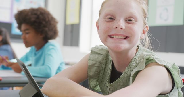 A young girl in a green dress is smiling while sitting at her desk in a classroom. She is looking directly at the camera with a cheerful expression. There is a blurred background showing another student. Ideal for use in educational materials, school advertising, and resources aimed at children’s education and development. Highlights themes of happiness, concentration, and technology in learning.