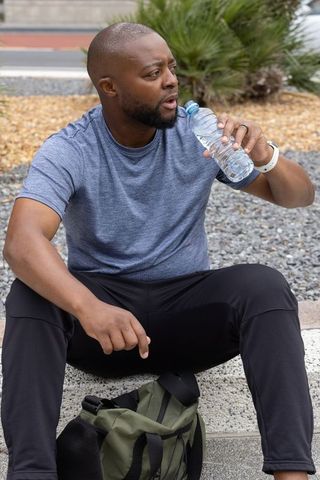 African american man taking water break after outdoor run sitting on curb with backpack
