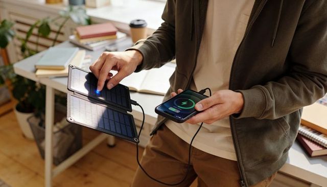 Man charging smartphone with foldable solar panel at home desk, sustainable portable power