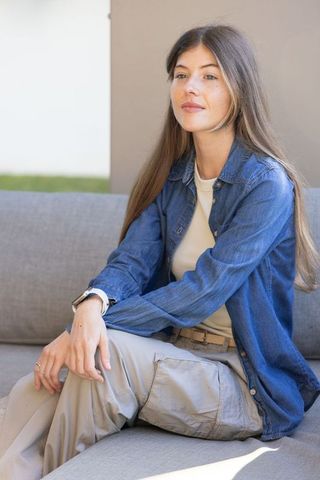 Young woman sitting on sunny patio sofa wearing denim shirt and beige cargo pants, contemplative