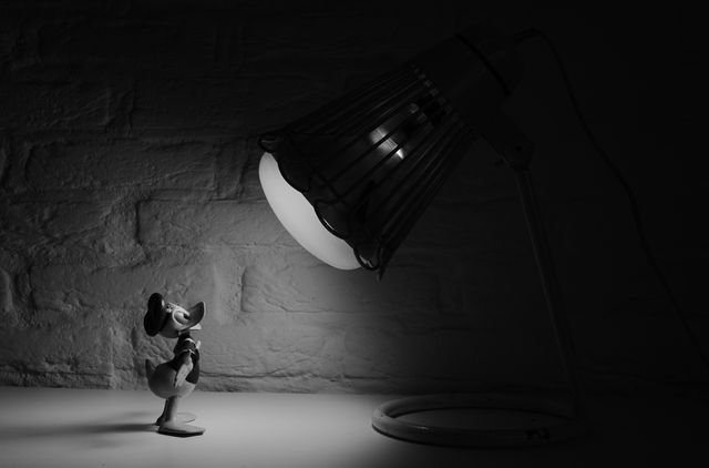 Classic Donald Duck toy standing under desk lamp's illumination casts dramatic contrast. Ideal for use in articles, children’s content, nostalgic pieces, or decorative purposes for a child’s room. The black and white tones add a vintage feel, suitable for modern minimalist designs.