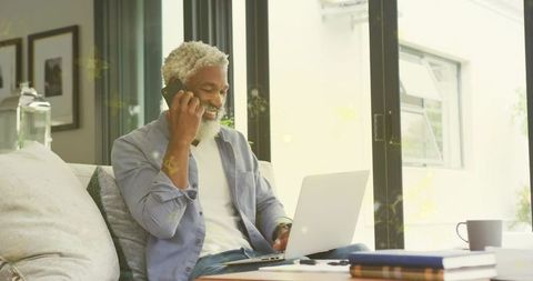 Senior man talking on phone while working on laptop in bright living room
