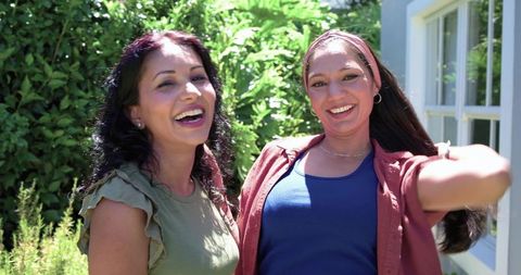 Two women laughing and posing in sunlit backyard with greenery and warm casual summer fashion