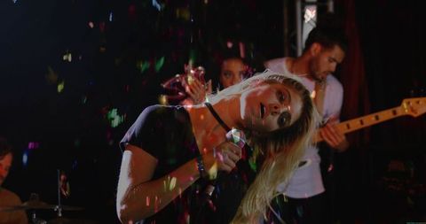 Young woman singing and leaning into mic under club lights with guitarist and confetti