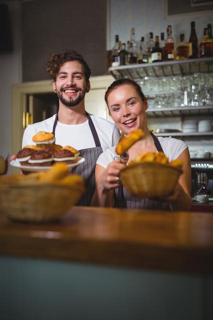Smiling waiter and waitress holding fresh baked goods in a cozy cafe. Ideal for use in marketing materials for cafes, bakeries, or restaurants, as well as in articles or advertisements focusing on hospitality, teamwork, and customer service.