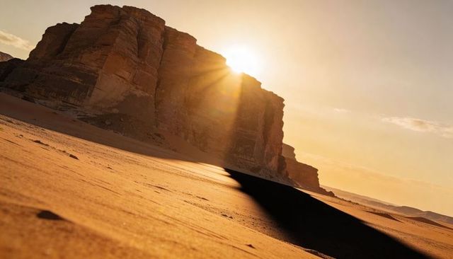 Sunburst lighting sandstone mesa casting long diagonal shadow across golden desert dunes